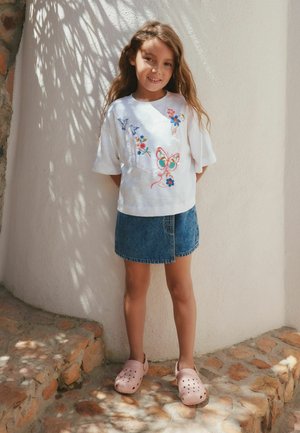 Young girl with long hair stands against white wall, wearing a white butterfly and flowers embroidered shirt, denim skirt, and pink slip-on shoes.