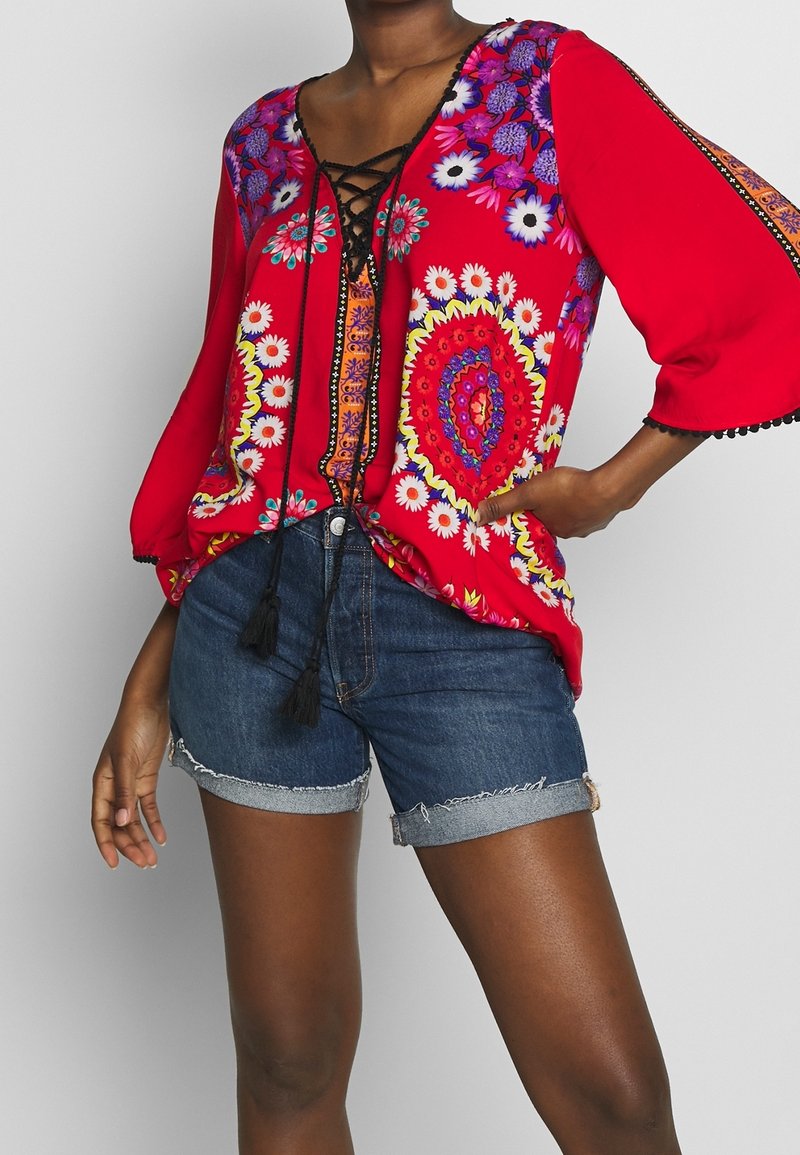 Blusa rossa a fiori con maniche a campana 3/4, scollatura con lacci e pompon neri; abbinata a pantaloni shorts in denim strappati con orli arrotolati.