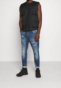 Chaleco sin mangas negro con múltiples bolsillos frontales, combinado con jeans azul desgastados y zapatillas negras. Diseño casual y funcional.