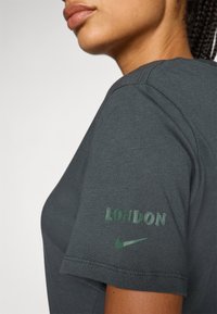 Grå bomullströja med texten "LONDON" och Nike-logotyp i grönt på ärmen. Designen har en avslappnad passform och en mjuk textur.
