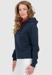 Felpa blu navy in tessuto morbido con cappuccio regolabile, polsini e orlo a costine. Indossata sopra una sciarpa rossa a pois e pantaloni bianchi.