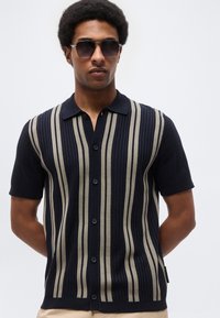 Άνδρας που φοράει σκούρα γυαλιά ηλίου και μπλε ναυτικού χρώματος κοντομάνικο πουκάμισο με κάθετες μπεζ ρίγες, στέκεται μπροστά σε ένα απλό φόντο.