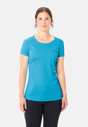 Donna in piedi, rivolta in avanti, che indossa una maglietta blu a maniche corte e pantaloni neri contro uno sfondo chiaro uniforme.