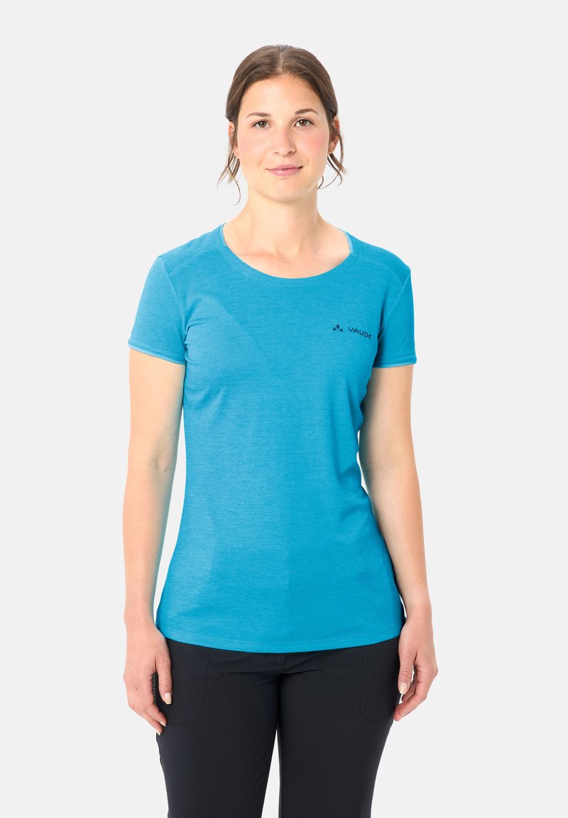 Donna in piedi, rivolta in avanti, che indossa una maglietta blu a maniche corte e pantaloni neri contro uno sfondo chiaro uniforme.