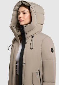 Beige puffer jacket with a large hood, black zipper, adjustable drawstrings, and a logo patch on the left sleeve. Soft, insulated texture.