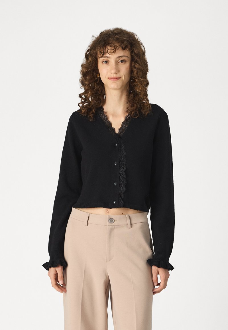 Melnā īsā kardigāna ar mežģīņu apdari, pogu aizdari un garām piedurknēm ar frillēm, saskaņota ar beizha augsto vidukļa biksēm.