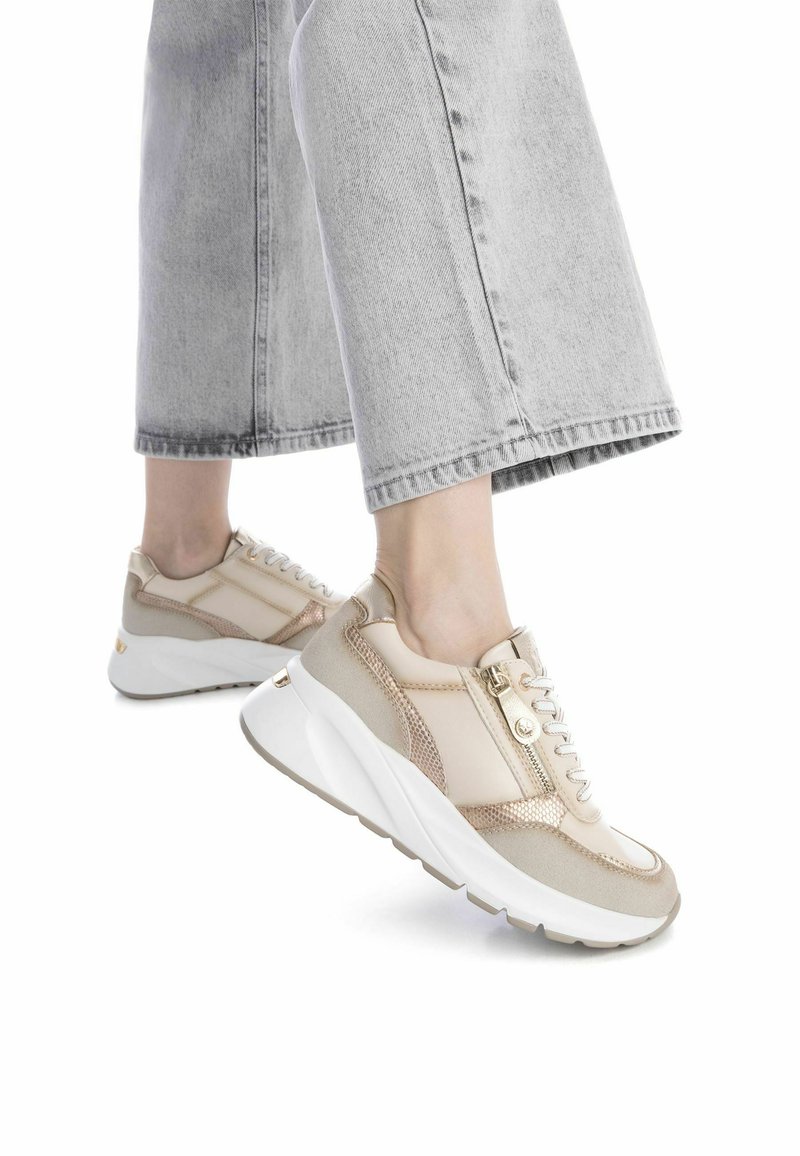 Zapatillas beige con acentos metálicos, detalles de malla y una suela blanca gruesa. Presentan un cierre de cremallera y cordones para un ajuste seguro.