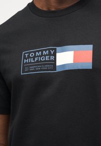 Μαύρο βαμβακερό T-shirt με ορθογώνιο λογότυπο που γράφει "TOMMY HILFIGER" σε λευκό, με μπλε φόντο και οριζόντιες κόκκινες και λευκές λωρίδες.