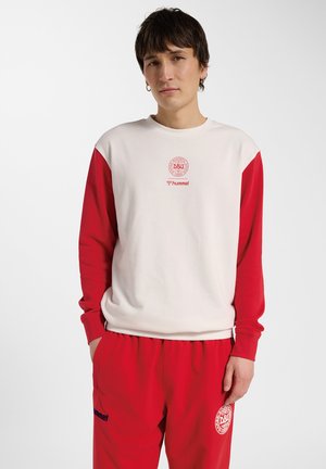 Ung person iført hvid sweatshirt med røde ærmer og røde bukser, med DBU og hummel sportslogoer, står med hænderne i lommerne.