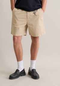 Beige shorts made of cotton, featuring side pockets and a drawstring waist. Paired with white socks and black loafers.