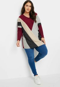 Top de manga larga en V de color rojo y gris con patrón de chevron, combinado con jeans skinny azules y zapatillas blancas. Tejido suave y texturizado.