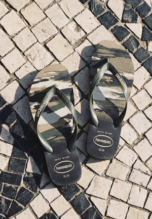 Par de chanclas Havaianas con patrón de camuflaje descansando sobre un pavimento de piedra en mosaico blanco y negro.
