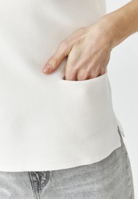 Valge tekstureeritud topp, millel on küljetasku, kus on käsi sees. Alumine äär on kergelt kaardus, complementides heledat halli kangast.