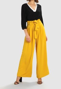 Top nero a scollo a V con bordino bianco, abbinato a pantaloni gialli a vita alta e a gamba larga con cintura a fiocco. La modella indossa tacchi neri.