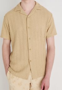 Korte mouwen, lichtbeige knoopshirt van een textuurmet een patroon. Beschikt over een spread collar en vijf knopen aan de voorkant.