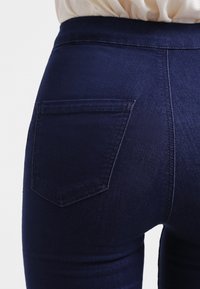 Tmavě modré džínové kalhoty s hladkým povrchem, které mají jedno zadní kapsu a vysoký pas, viditelné prošívání podél švů.
