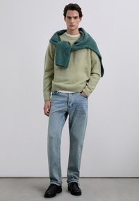 Suéter de lana verde claro con puños acanalados y una bufanda verde oscuro sobre los hombros, combinado con jeans azul claro y zapatos negros.