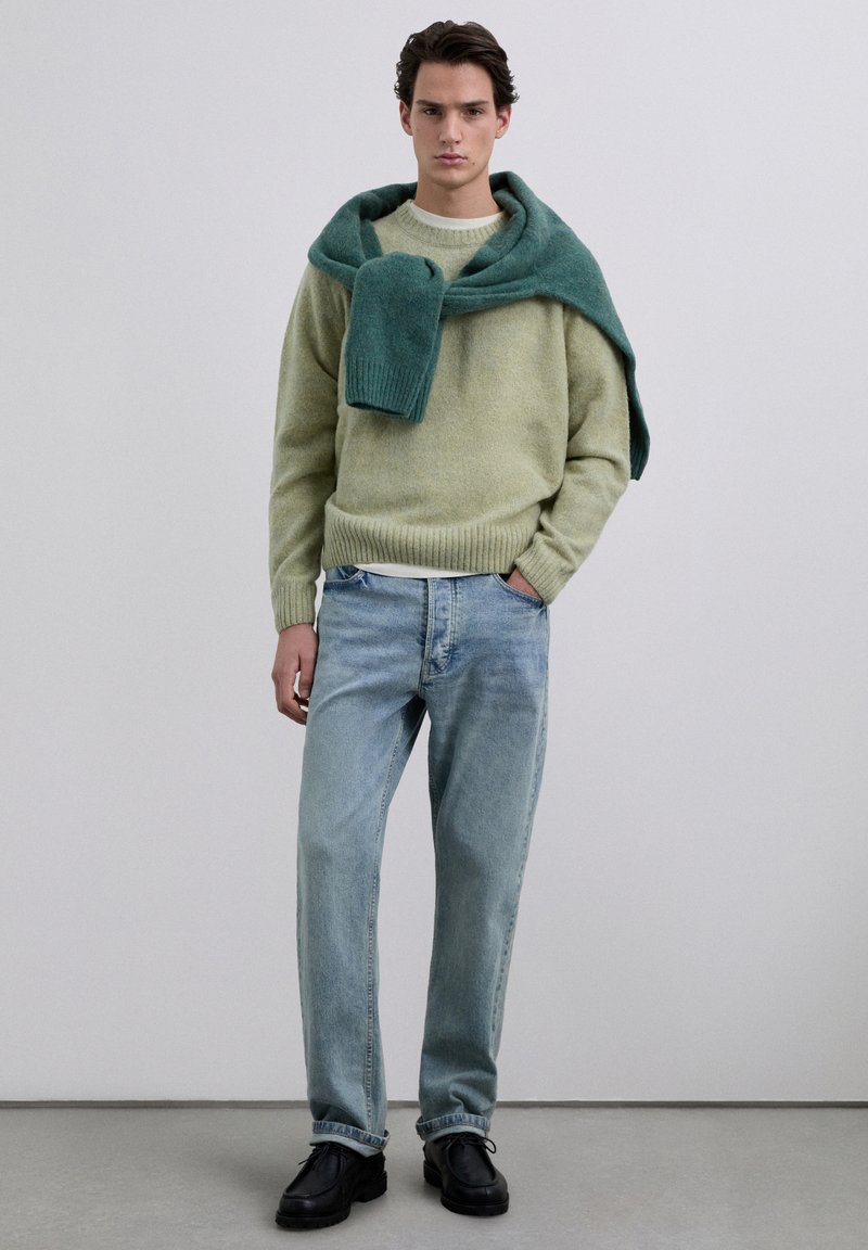 Suéter de lana verde claro con puños acanalados y una bufanda verde oscuro sobre los hombros, combinado con jeans azul claro y zapatos negros.