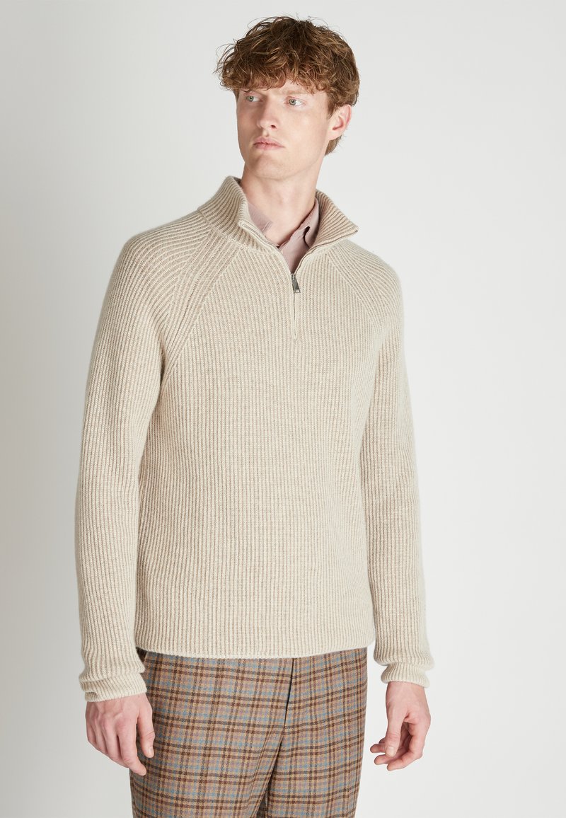 Beige gerippter Strickpullover mit Halbreißverschluss und Raglanärmeln. Weiche Textur, hoher Kragen und ein Reißverschlussdetail am Hals. Kombiniert mit karierter Hose.