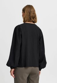 Black textured blouse with long, slightly puffed sleeves. Features vertical ribbing and a round neckline, paired with light gray trousers.