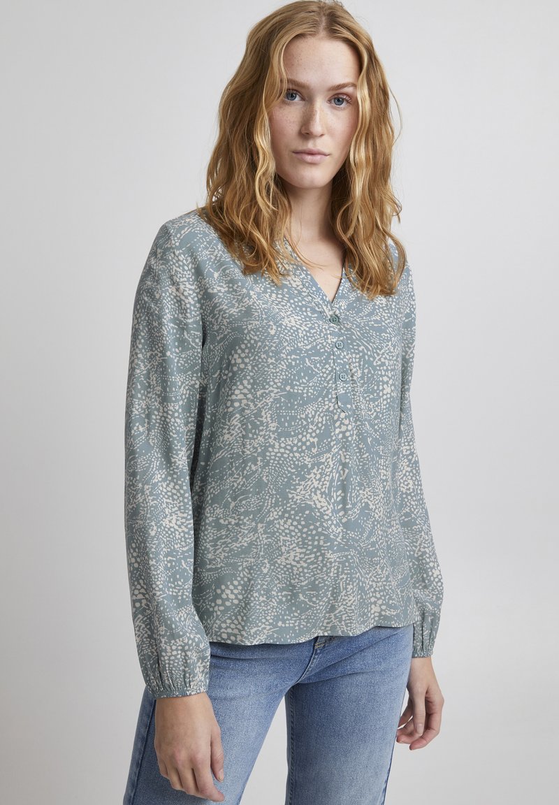 Camicia blu chiaro con bottoni, decorata con un motivo floreale bianco. Presenta maniche lunghe con polsini arricciati e un design a V. Tessuto morbido.