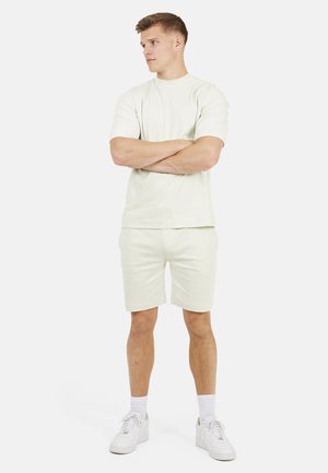 Młody mężczyzna stoi ze skrzyżowanymi ramionami, ubrany w kremową koszulkę, dopasowane szorty, białe skarpetki i białe trampki na jednolitym tle.