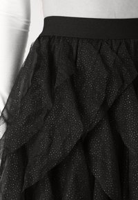 Jupe en tulle noire à plusieurs couches avec de petits pois argentés et une large ceinture noire portée avec un top blanc ajusté à manches longues.