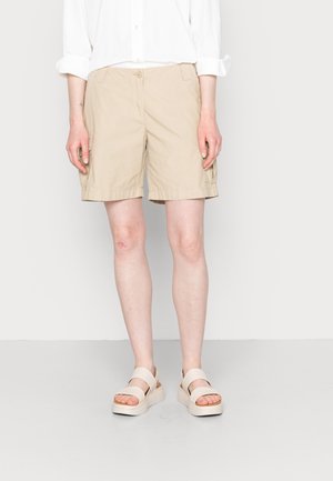Person trägt beige kniehohe Shorts, weißes hochgekrempeltes Langarmhemd und beige Plateau-Sandalen, steht vor weißem Hintergrund.