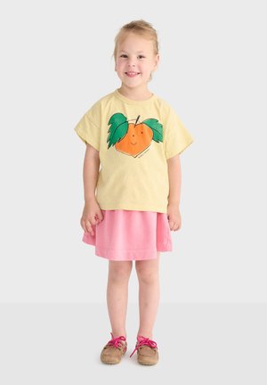 Jeune fille souriante portant un t-shirt jaune avec un motif pêche, une jupe rose, et des chaussures marron aux lacets roses sur un fond uni.