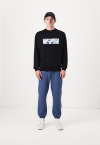 Sudadera negra con un parche de patrones coloridos, combinada con pantalones de chándal azules y zapatillas blancas. El modelo lleva una gorra negra.