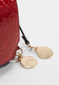 Esquina de bolso rojo texturizado con dos etiquetas redondas doradas grabadas con "RIVER ISLAND," unidas por anillos dorados a una correa negra.