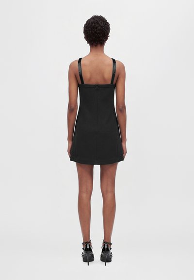 Un mannequin se tient dos à la caméra, vêtu d’une mini-robe noire sans manches et de chaussures noires à talons hauts, sur un fond clair et uni.