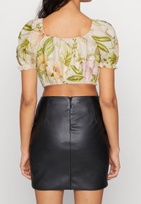 Femme portant un haut court à manches bouffantes fleuries et une mini-jupe en cuir noire taille haute, vue de dos sur un fond uni.