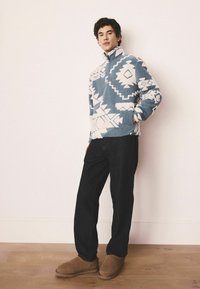 Blue and white patterned fleece pullover with a high collar, paired with dark denim pants and brown slip-on shoes. Wooden floor backdrop.