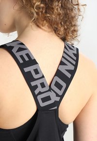 Černá sportovní podprsenka s širokými křížovými ramínky, na kterých je tištěný nápis "NIKE PRO" v šedé barvě. Hladká textura, přiléhavý design.