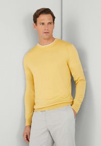 Suéter de lana amarillo con cuello redondo y mangas largas, que presenta un interior de color blanco en el cuello, combinado con pantalones de color gris claro.
