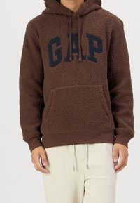 Hnědá mikina z fleecu s kapsou klokanem, s tmavě modrým logem "GAP" na hrudi a kapucí se šňůrkou. Měkká, strukturovaná látka.