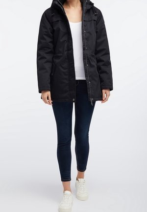 Femme portant une veste matelassée noire sur un haut blanc, un jean skinny foncé et des baskets blanches, debout devant un fond uni.