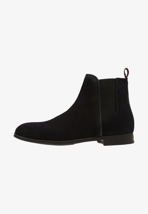 Bottine Chelsea en daim noir avec panneaux latéraux élastiques, talon bas et languette de tirage rouge à l'arrière.