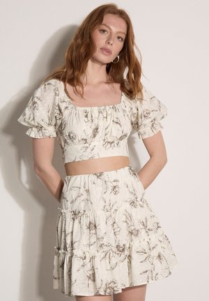 Kobieta o rudych włosach ubrana w biały kwiecisty dwuczęściowy strój z bufkami na krótkich rękawach i warstwową spódnicą, pozująca na jednolitym tle.