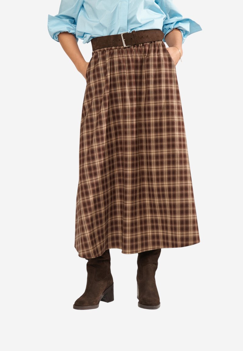 Jupe midi à carreaux bruns avec large ceinture, portée avec un chemisier bleu et des bottines à talons en daim brun foncé. Mains dans les poches.