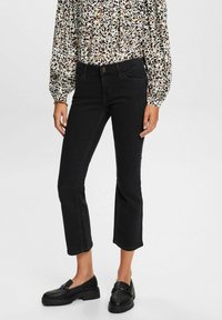 Mujer vistiendo vaqueros negros recortados, mocasines negros y una blusa de manga larga blanca con manchas abstractas negras y marrones.