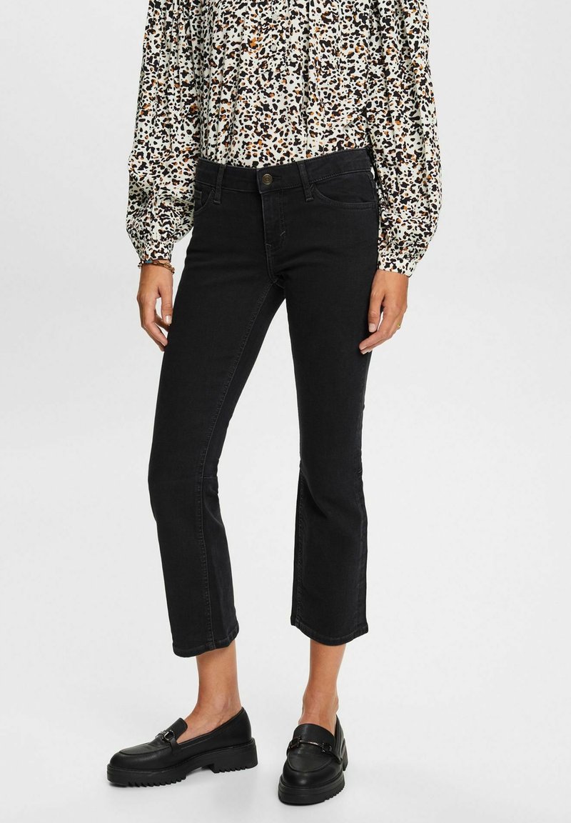 Mujer vistiendo vaqueros negros recortados, mocasines negros y una blusa de manga larga blanca con manchas abstractas negras y marrones.