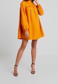 Oranssi, pitkähihainen, korkeakauluksinen mekko, jossa on rento leikkaus. Mekossa on rypytetty yksityiskohta rinnan alapuolella ja se on valmistettu sileästä, kevyestä kankaasta.