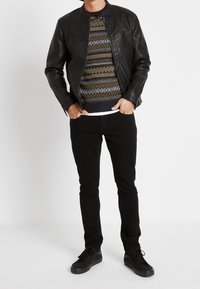 Svart läderjacka över en mönstrad tröja med geometriska mönster i grönt och blått, kombinerat med svarta byxor och svarta sneakers.