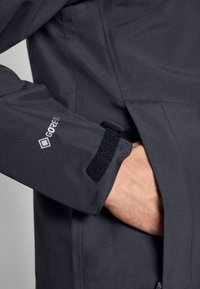 Chaqueta Gore-Tex en gris oscuro con una superficie texturizada. Cuenta con un cierre de puño de Velcro y un bolsillo oculto en el lateral.