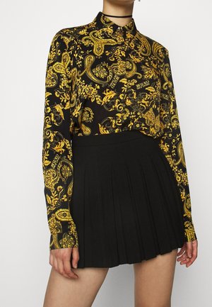 Kobieta ubrana w czarną bluzkę z długim rękawem z żółtymi wzorami paisley i czarną plisowaną mini spódnicę, z czarnym naszyjnikiem choker.