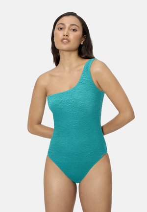 Mujer con cabello largo y oscuro que lleva un traje de baño turquesa texturizado de un solo hombro, de pie con los brazos detrás de la espalda sobre un fondo blanco.