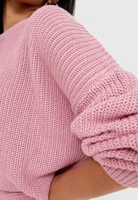 Gros plan sur une personne portant un pull rose tricoté texturé avec les manches retroussées, sur un fond clair uni.