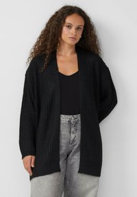 Czarny sweter z dzianiny z otwartym przodem, z oversize'owymi rękawami i teksturowanym wzorem. Noszony na czarnej bluzce i szarych dżinsach o wysokim stan.